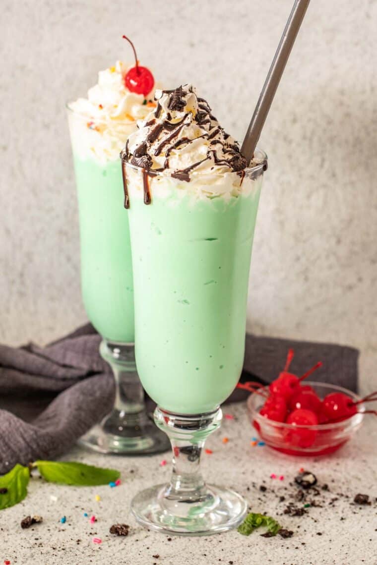 Shamrock shake with two glass cups topped with whipped cream, cherries and chocolate drizzle. 