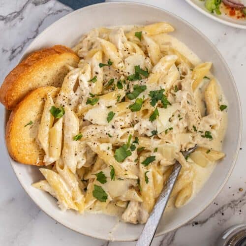 A bowl of chicken Alfredo with two slices of bread.