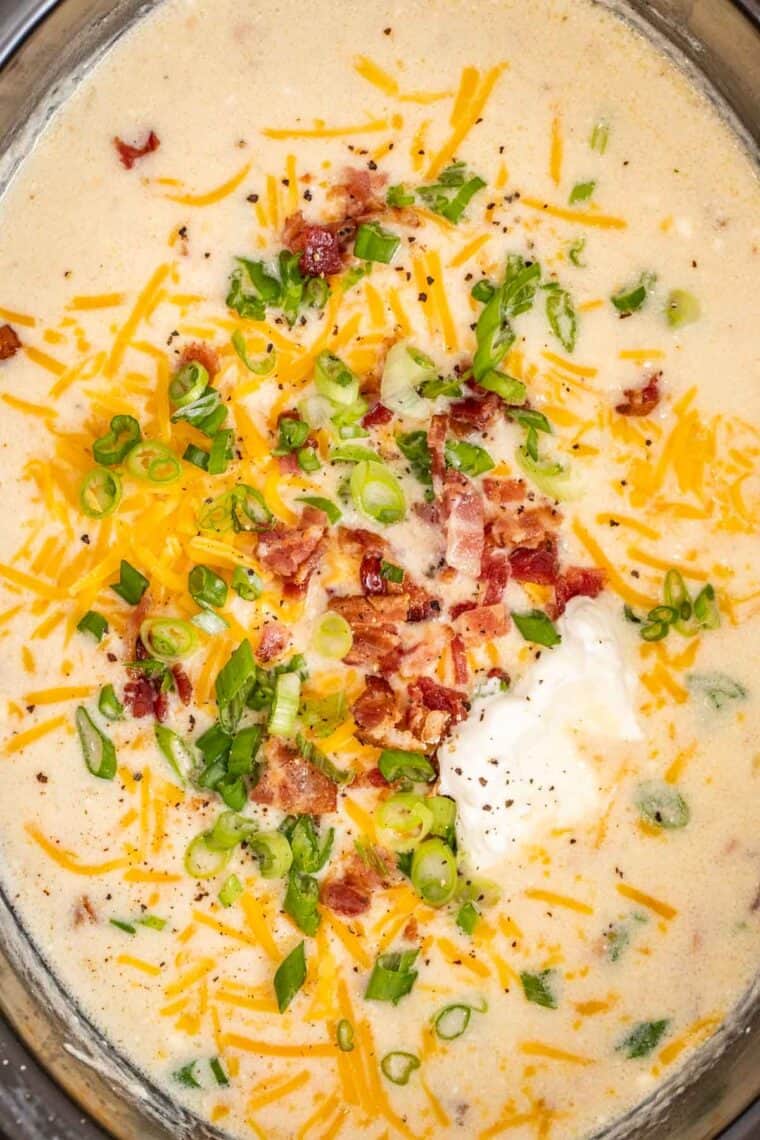 Creamy potato soup in a black slow cooker topped with sour cream, cheese, bacon and green onion.