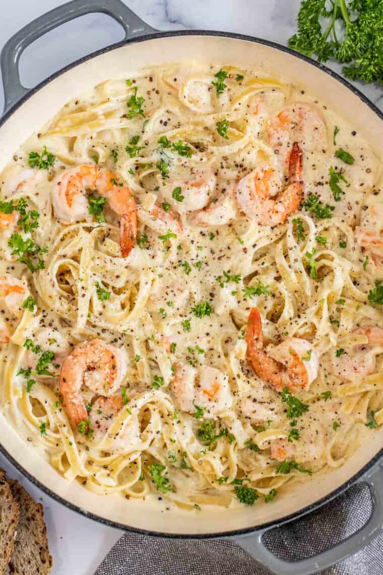 A deep skillet with shrimp Alfredo pasta topped with fresh parsley and ground black pepper. 
