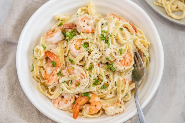 A white plate with creamy shrimp Alfredo pasta topped with greens. 
