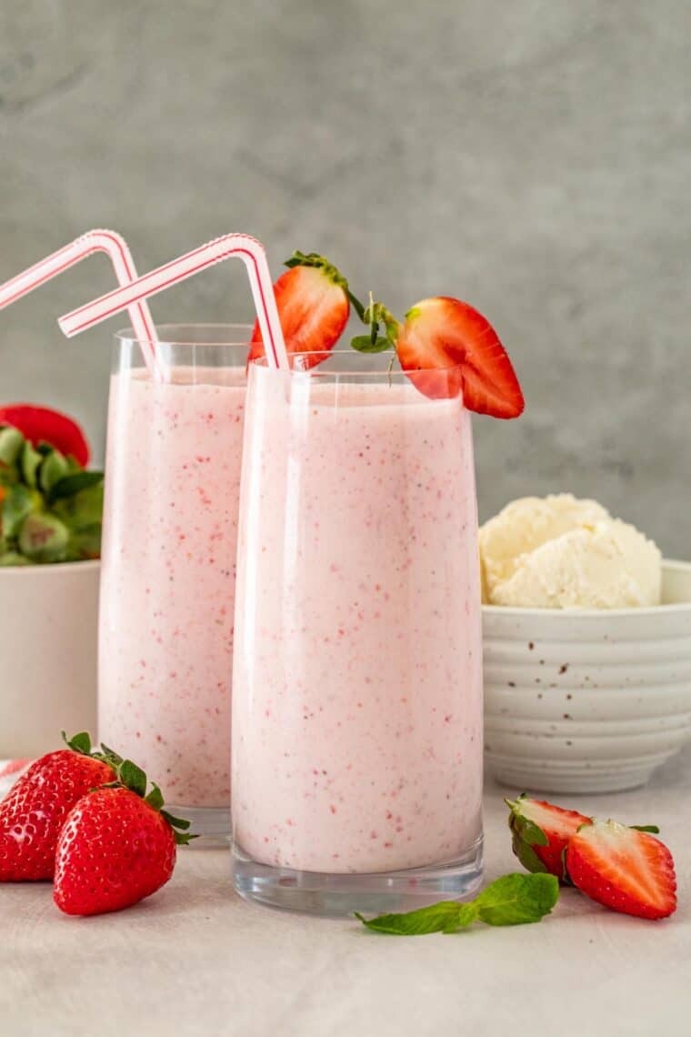 Strawberry milkshake in two glass cups topped with strawberries and straws. 