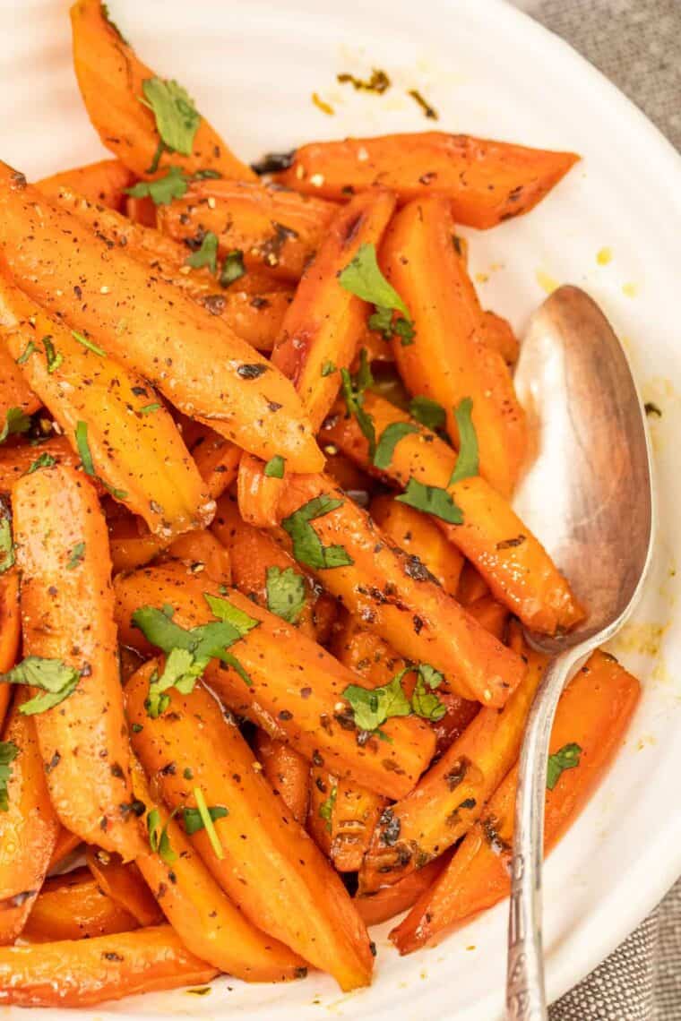 A white bowl of honey roasted carrots topped with fresh greens with a metal spoon. 