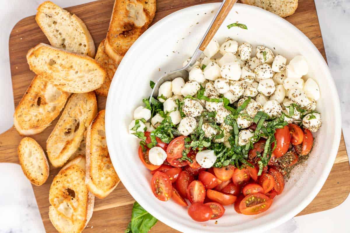 A bowl of caprese salad next to crostini  on a cutting board. 