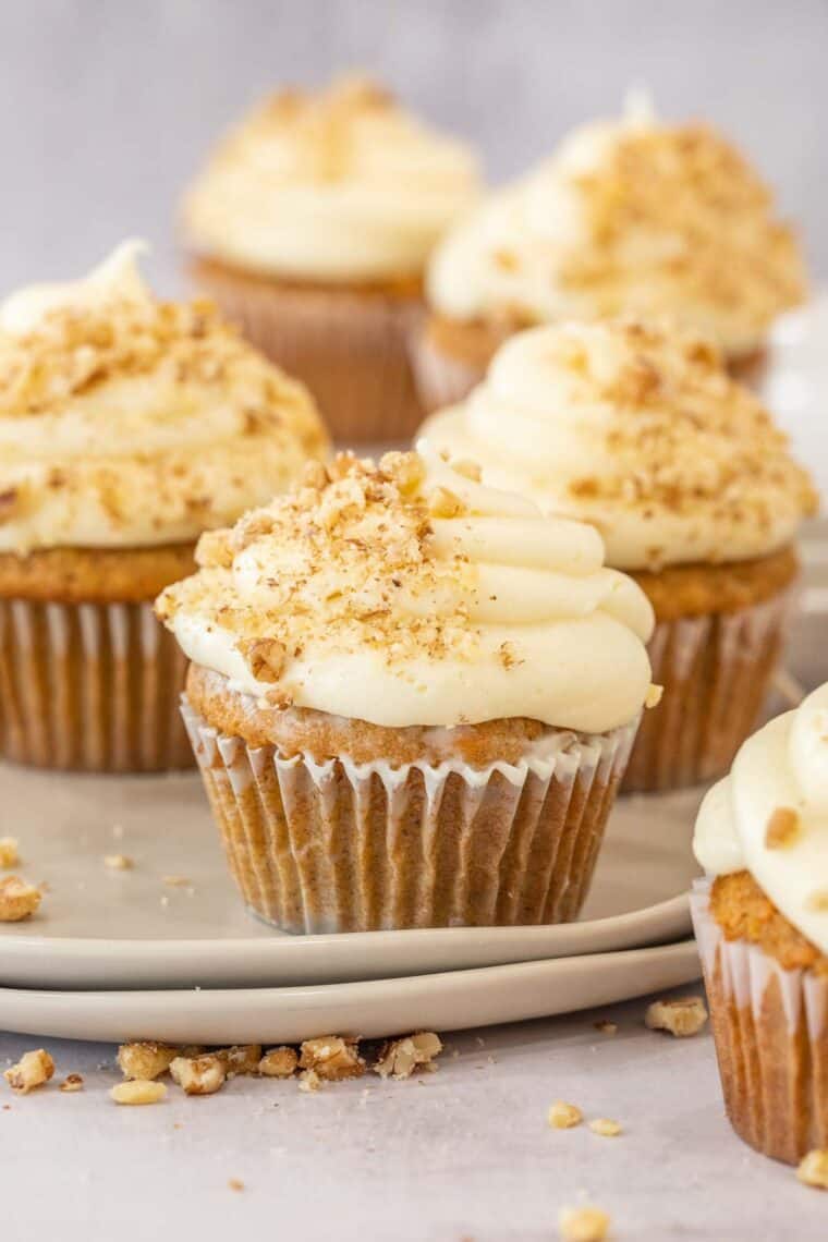 Carrot cupcakes on a tan plate topped with icing and chopped walnuts. 