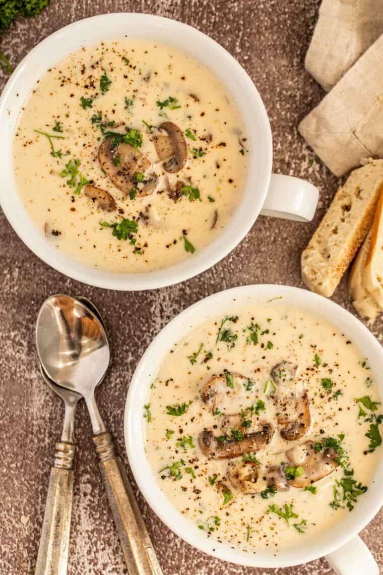 Two bowls of mushroom soup next to metal spoons and bread. 