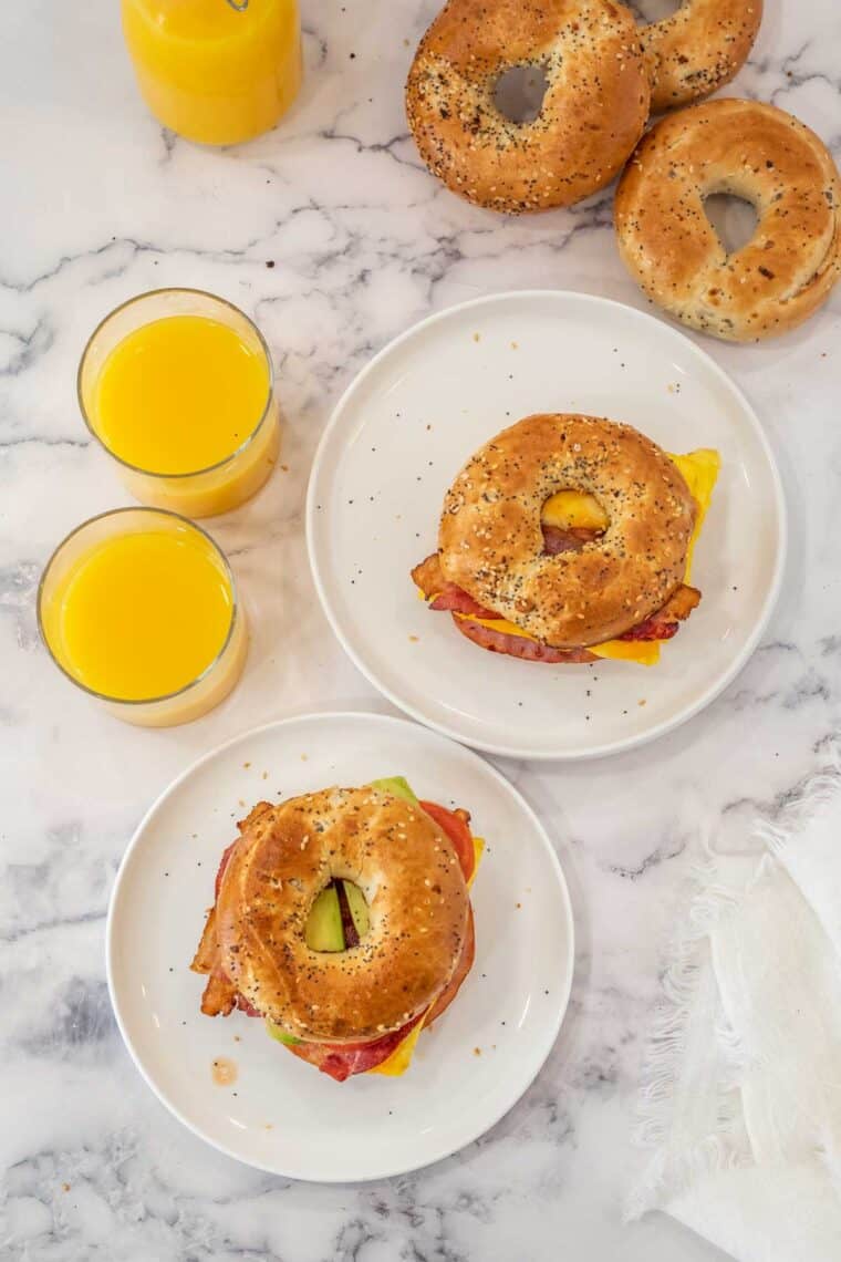 Two bagel sandwiches on white plates next to glasses of orange juice. 