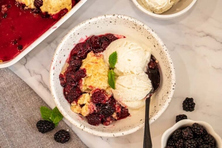 Blackberry cobbler in a white bowl with ice cream and a spoon. 