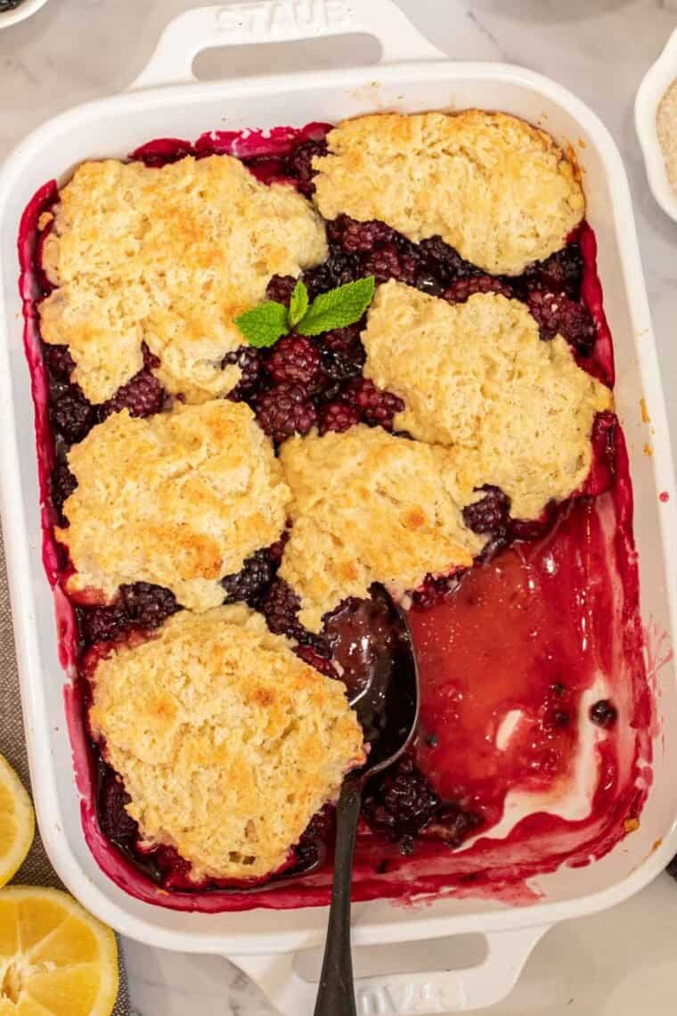 A white casserole dish with blackberry cobbler and a black spoon.