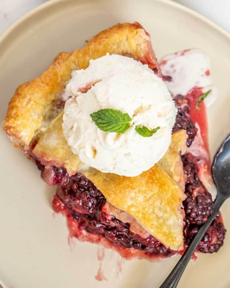 A slice of balckberry pie topped with vanilla ice cream with a black spoon. 