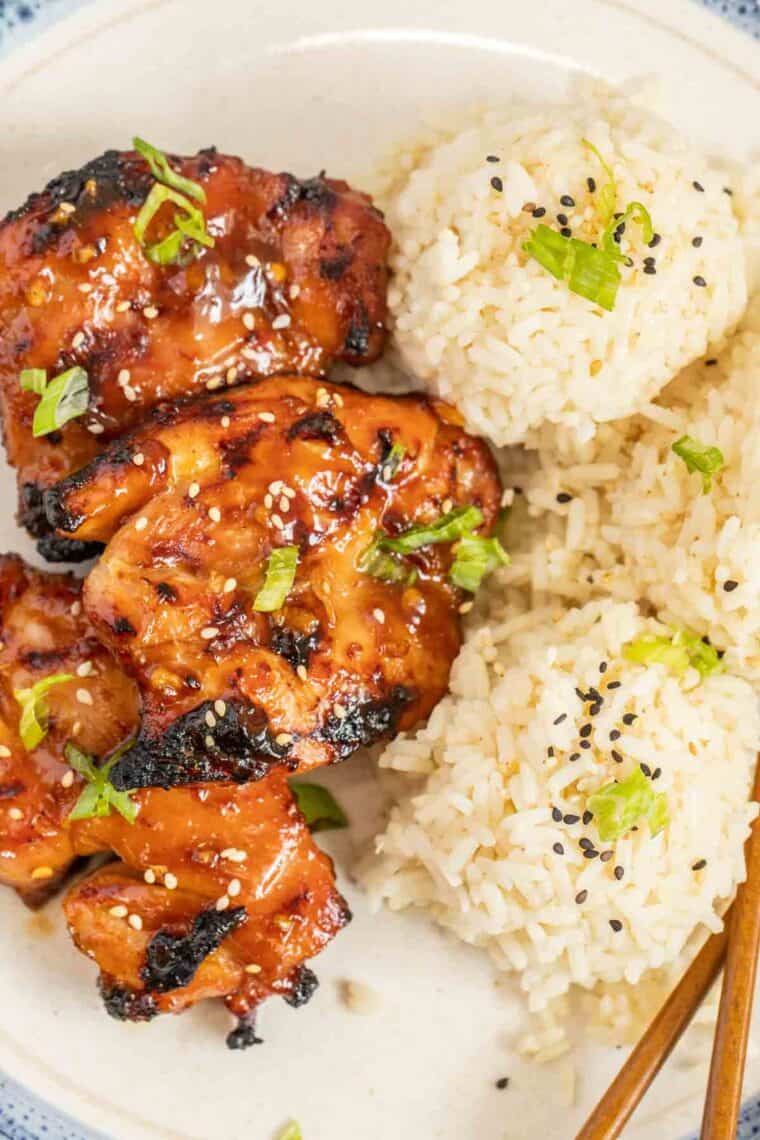 Grilled teriyaki chicken on a plate with rice topped with green onion ad sesame seeds. 
