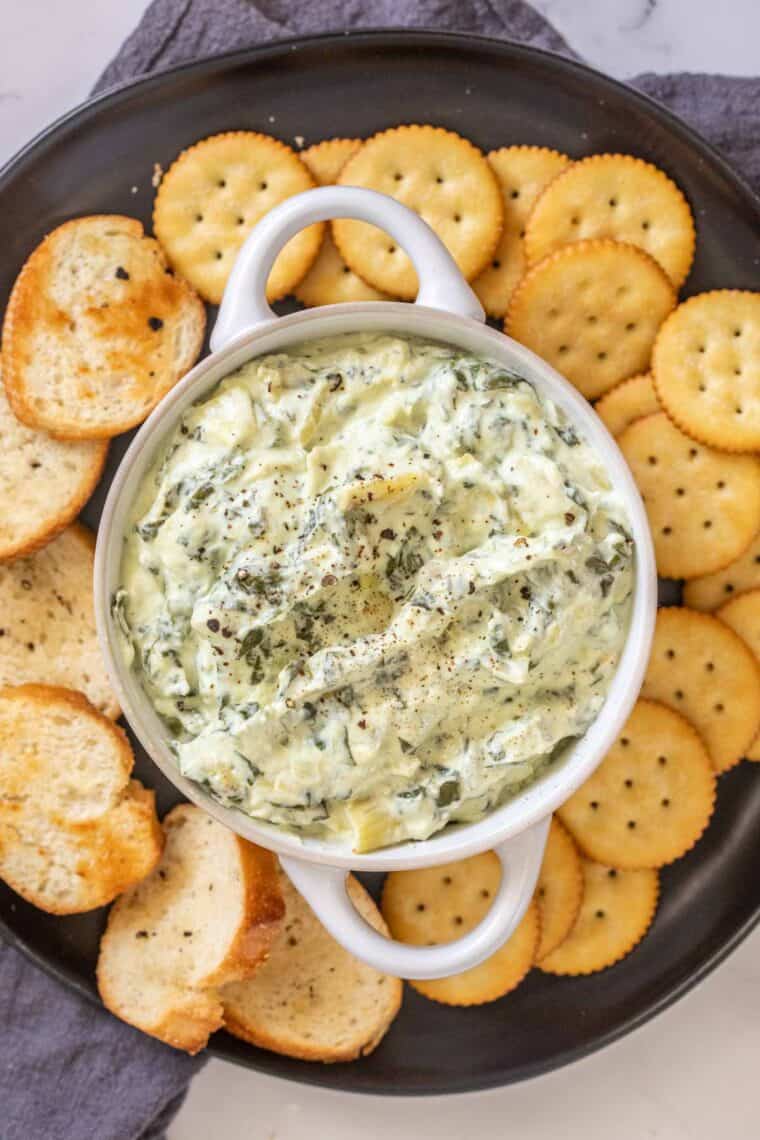 A black plate with crackers in a white creamy spinach artichoke dip. 