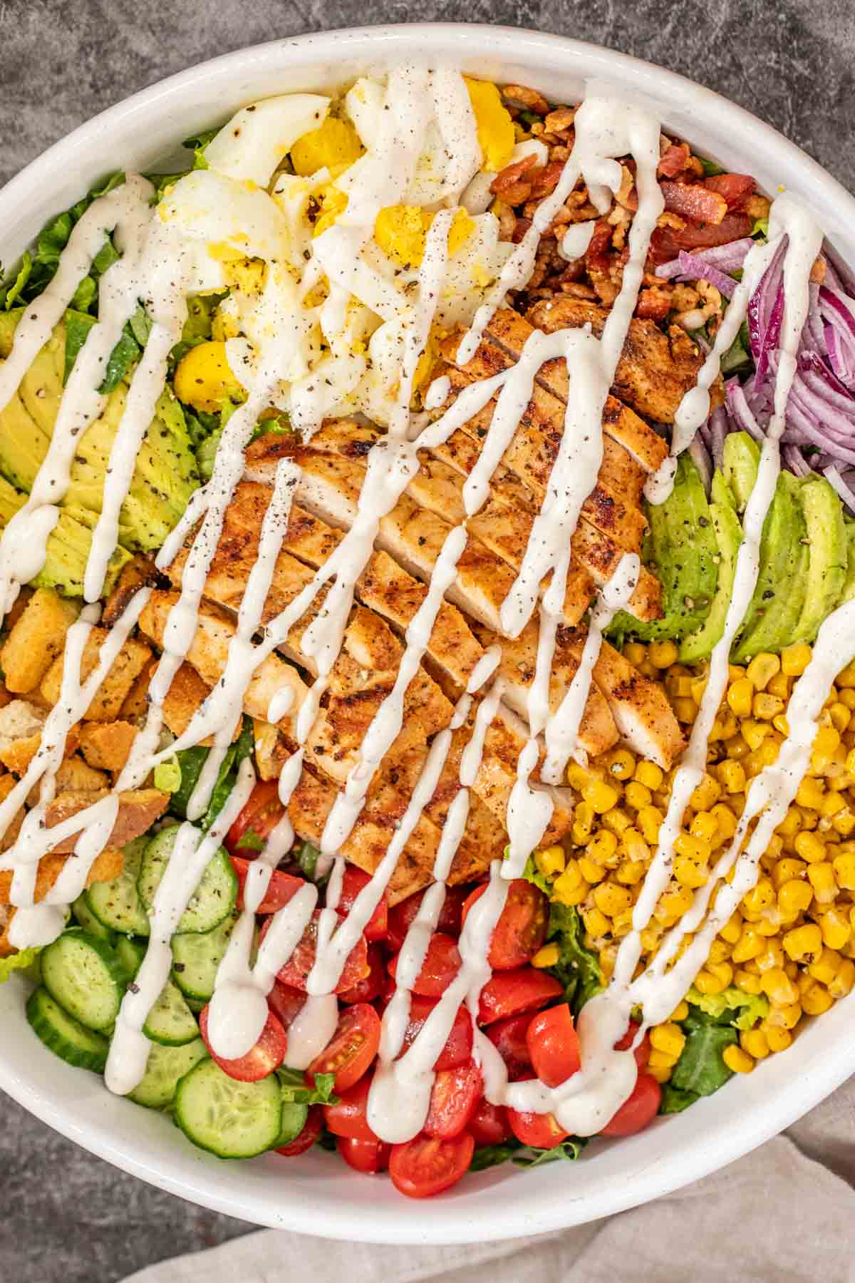 A salad bowl with the grilled chicken along with all the salad ingredients. The top is drizzled with ranch dressing.