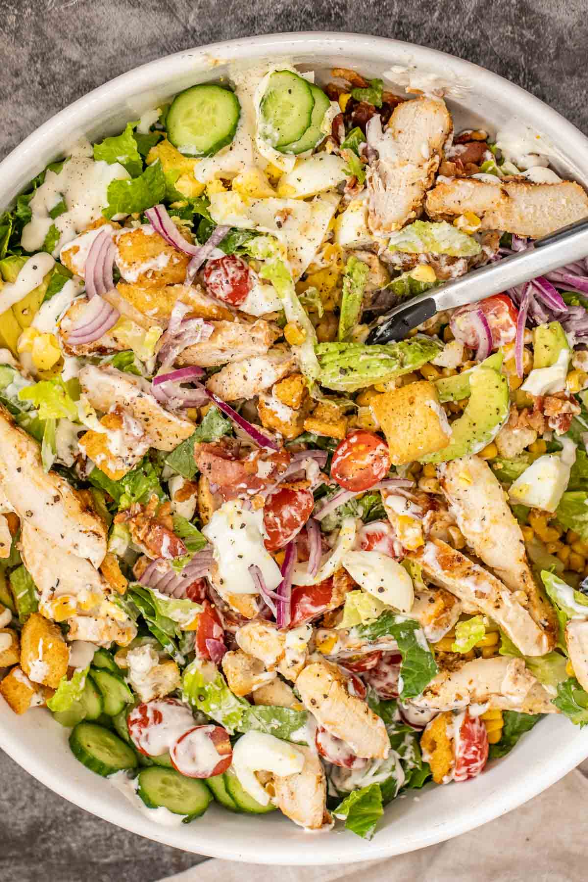 There mixed grilled chicken salad along with a pair of tongs.