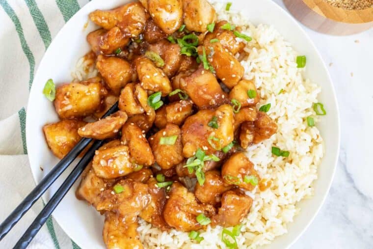 A white plate of rice and orange chicken topped with green onion. 