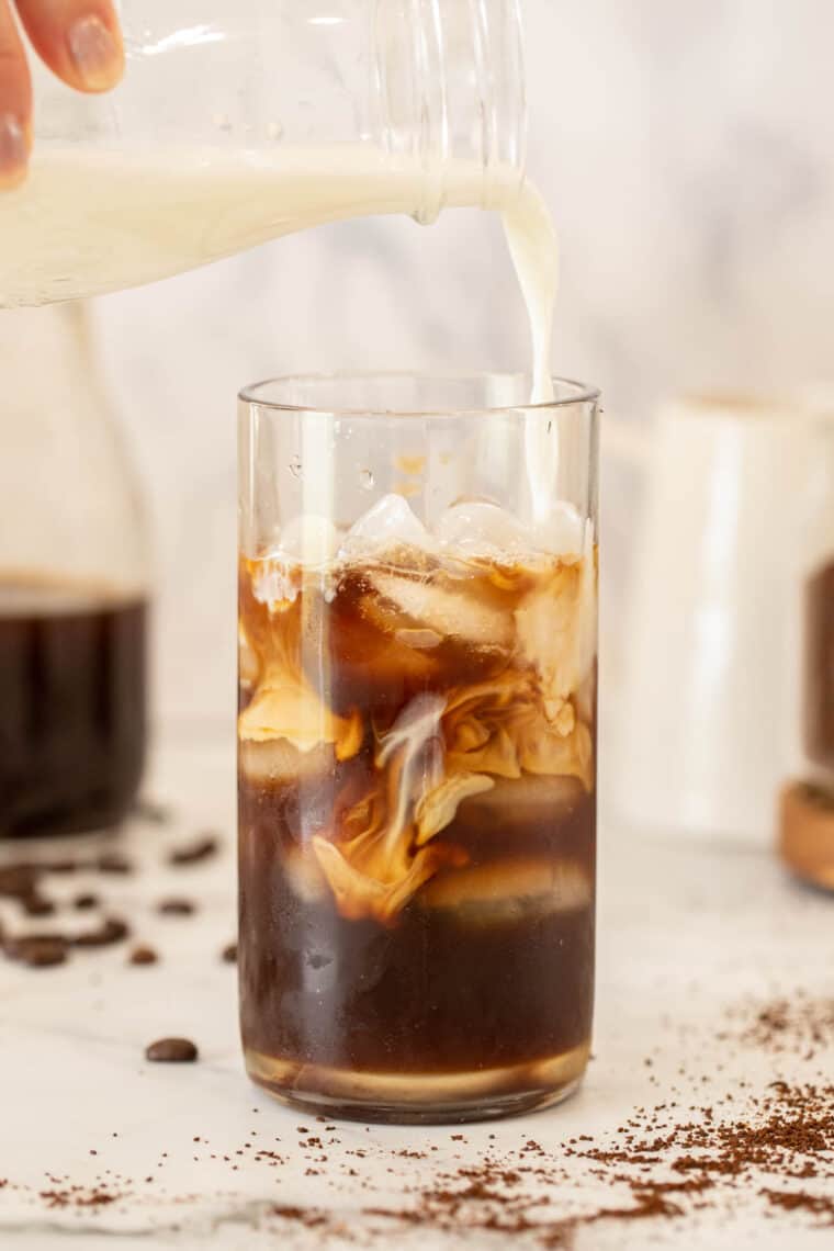 Cold brew coffee in a glass over ice with creamer being poured into the glass. 