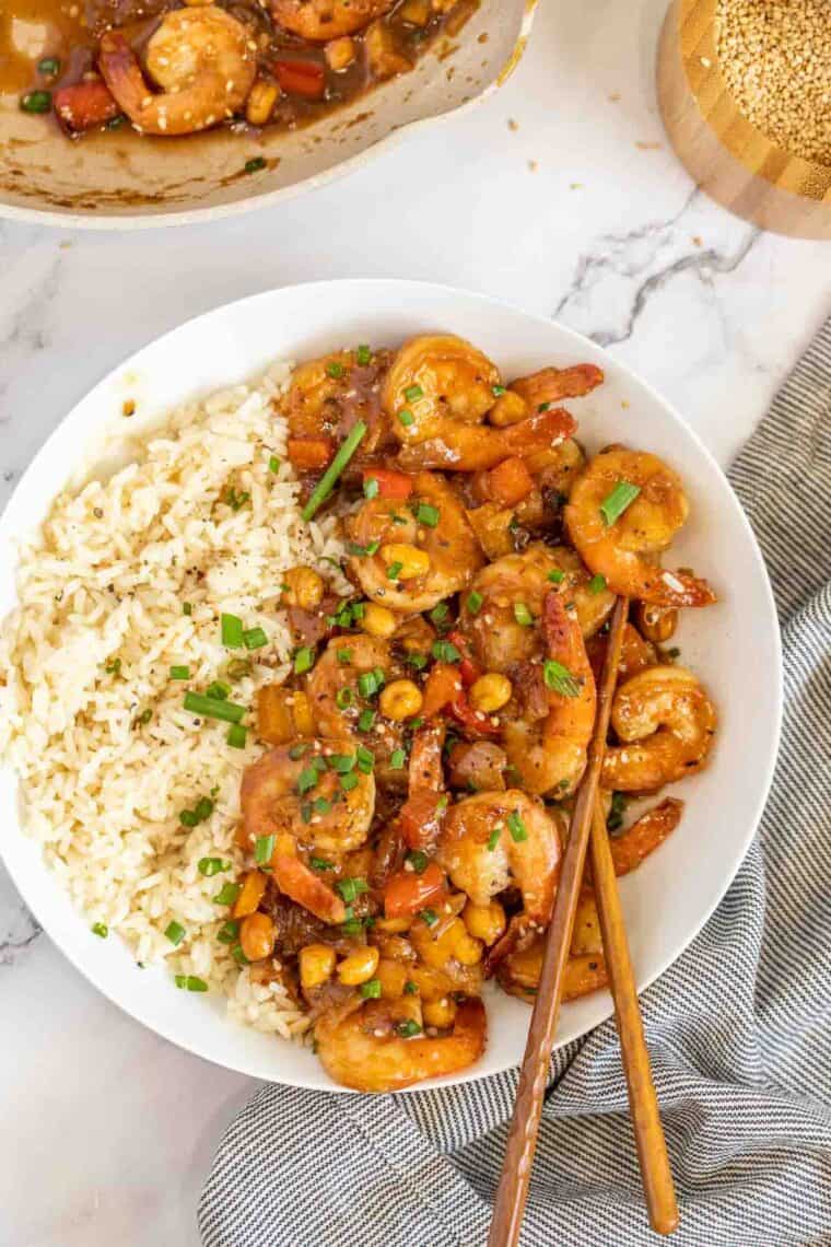 Kung Pao shrimp in a white plate with rice topped with fresh greens. 