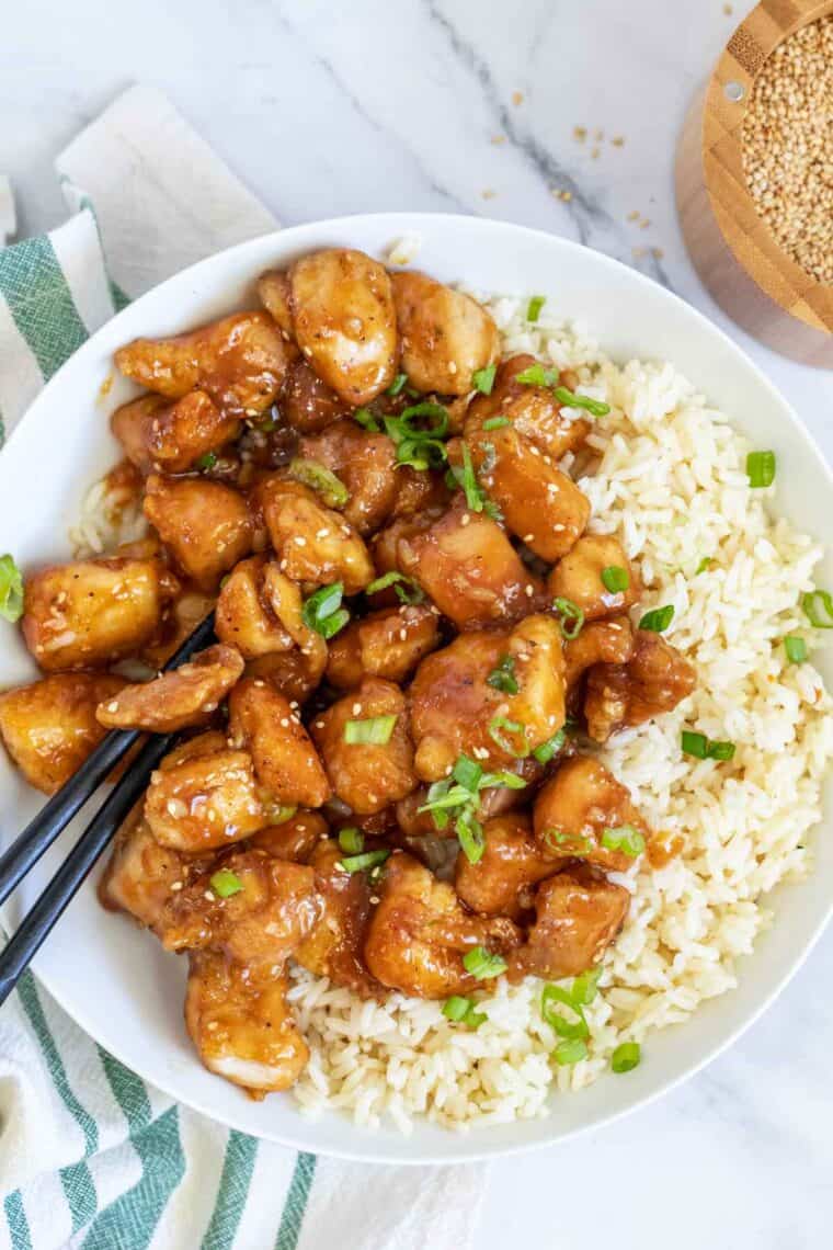 A white bowl with rice topped with homemade orange chicken topped with fresh green onion. 