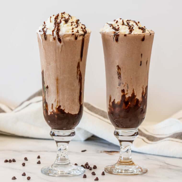 Two chocolate milkshakes with whipped cream and chocolate syrup.
