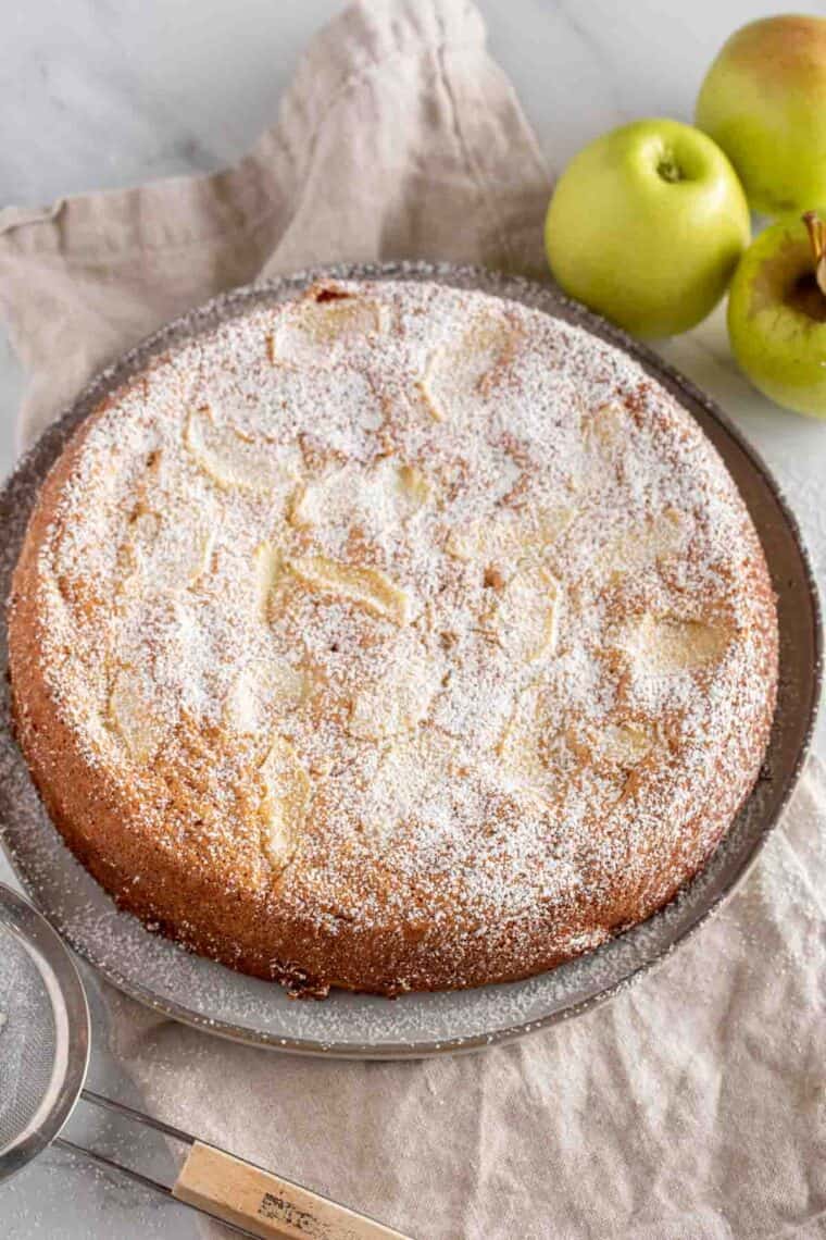 Apple cake served on a plate with apples and powdered sugar on the side.