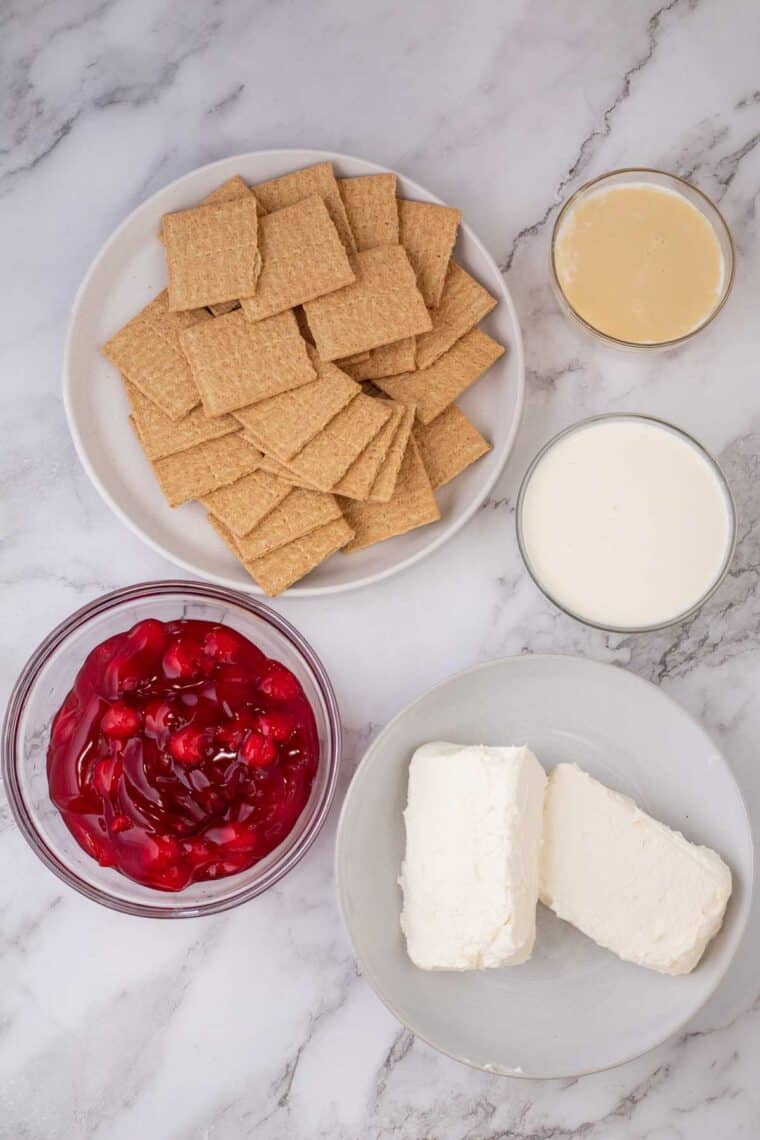 Ingredients needed to make cherry cheesecake next to eachother in separate plates and/or bowls.