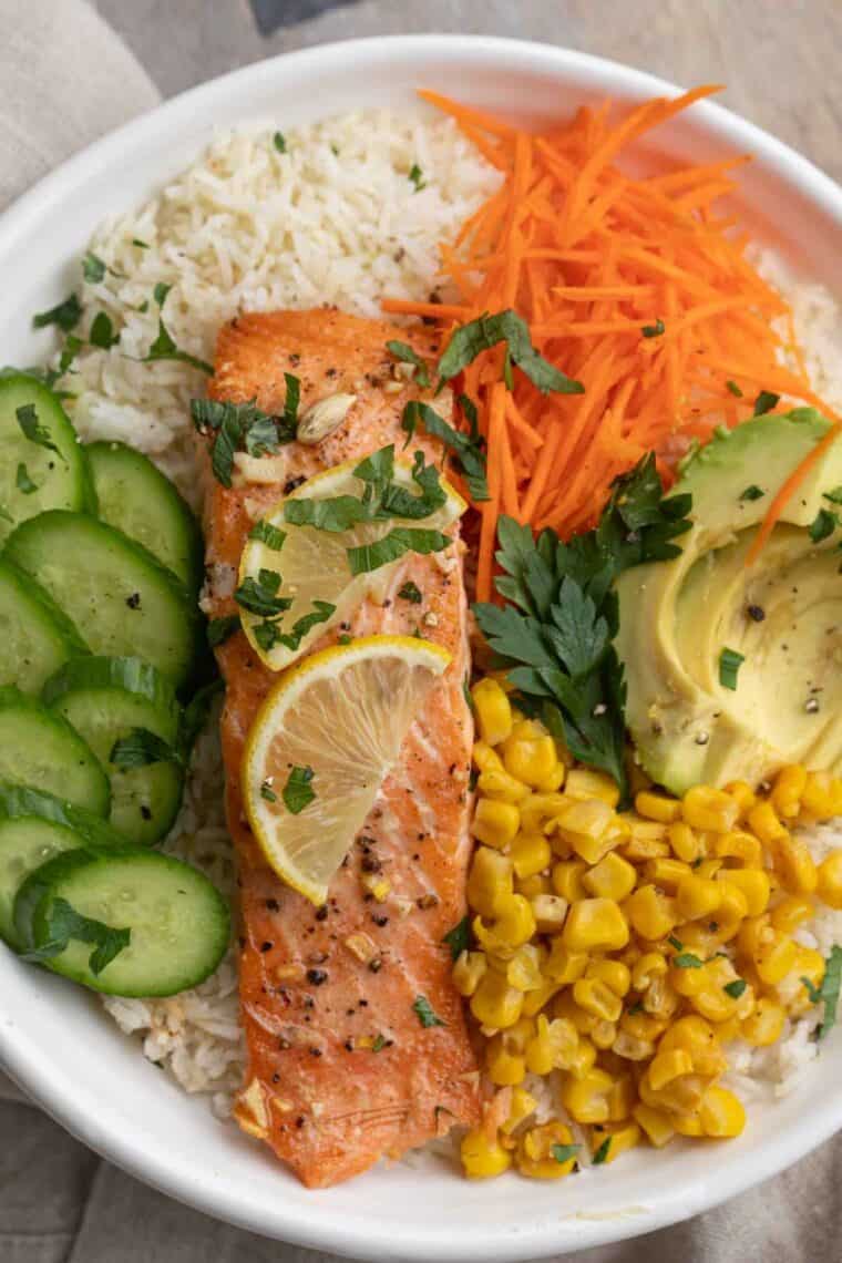 Close up of salmon bowl served with sliced avocados, sliced carrots, corn, sliced cucumbers, and lemon.
