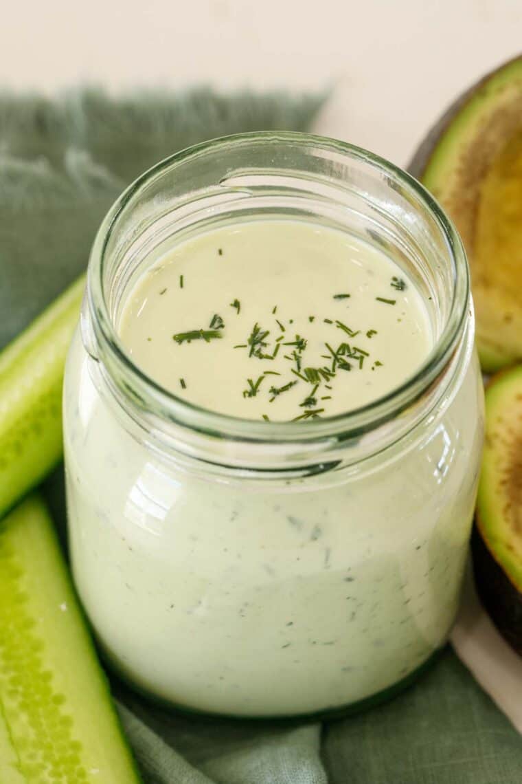 Close up of avocado ranch dressing in a jar with a side of cucumbers and topped with greens.