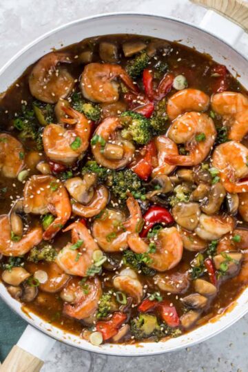 Shrimp stir fry in a pan with a spatula and sesame seeds in the background.