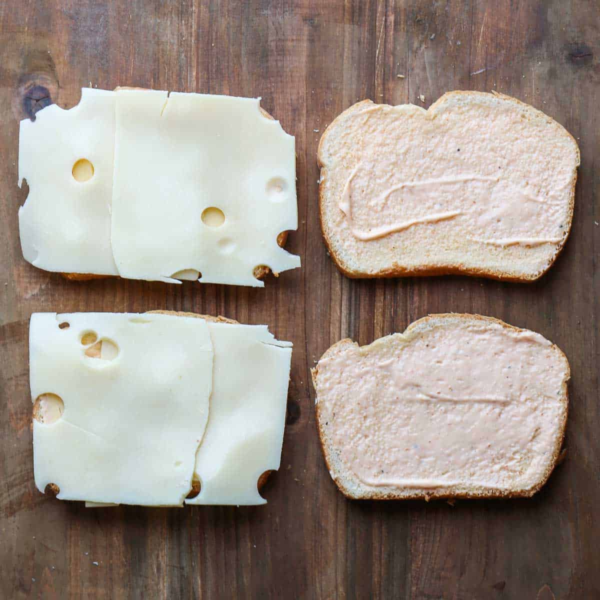 Four slices of bread laid out. Two slices of bread have a thin layer of sauce, while the other two slices have Swiss cheese.
