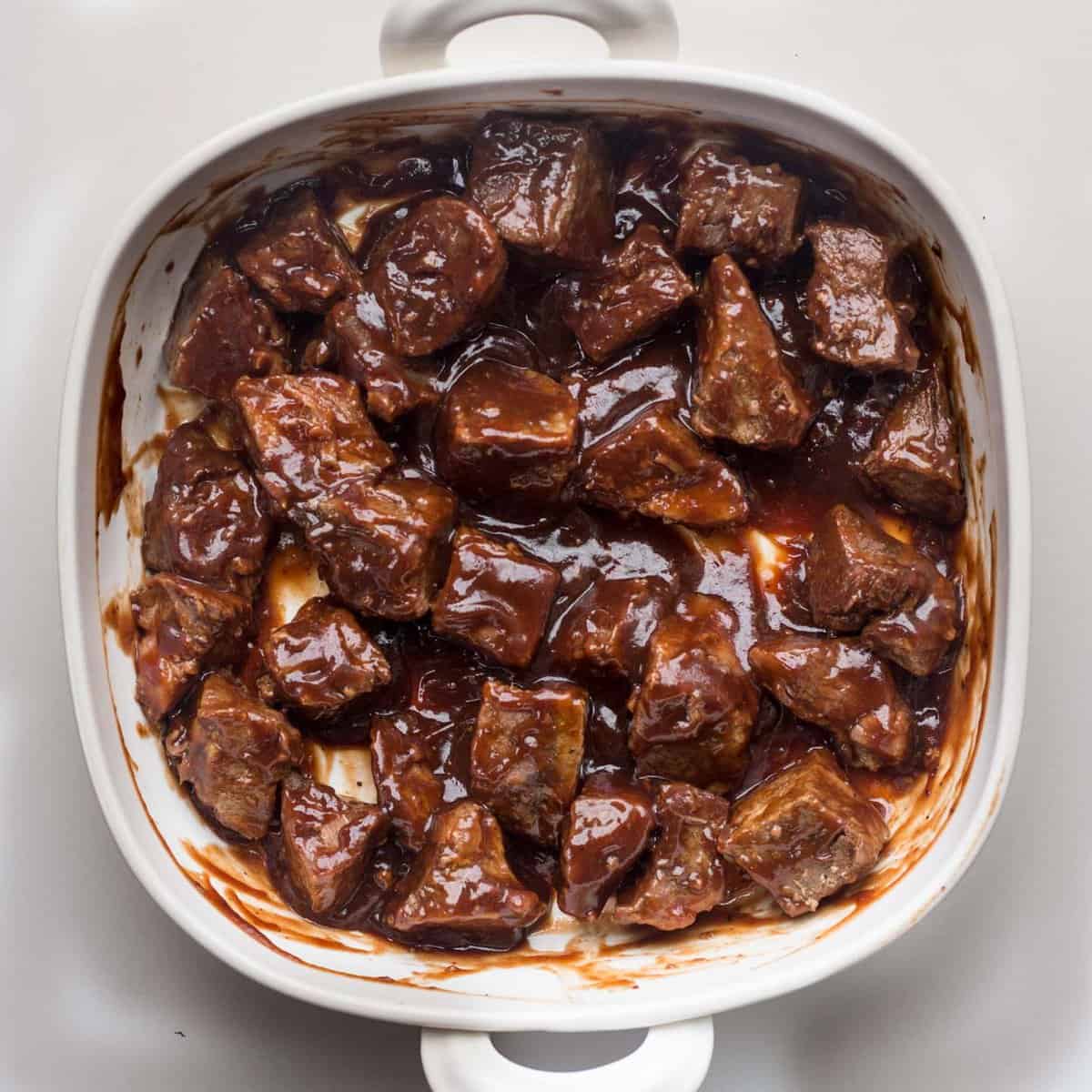 A white casserole dish containing all the beef chunk covered in BBQ sauce. 
