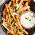 A black plate with parmesan fries with a garlic aioli dipping sauce.