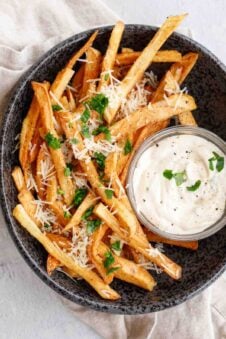 A black plate with parmesan fries with a garlic aioli dipping sauce.