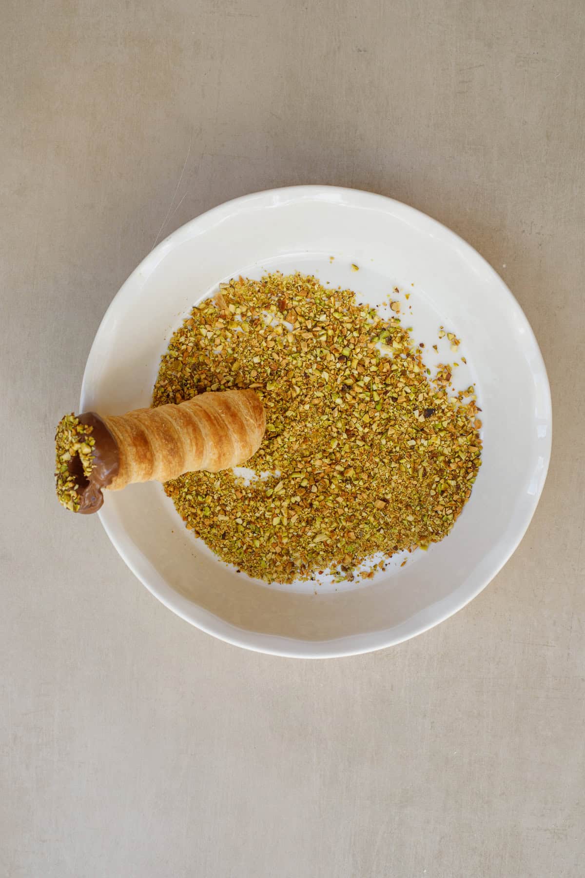 These easy to make dubai chocolate puff pastry horns make the perfect dessert. A plate with crushed pistachios and a chocolate coated horn dipped into it.