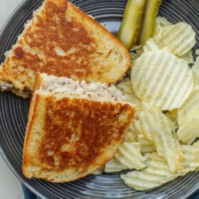 A plate with a cooked tuna melt sandwich, accompanied by crispy potato chips and pickles.