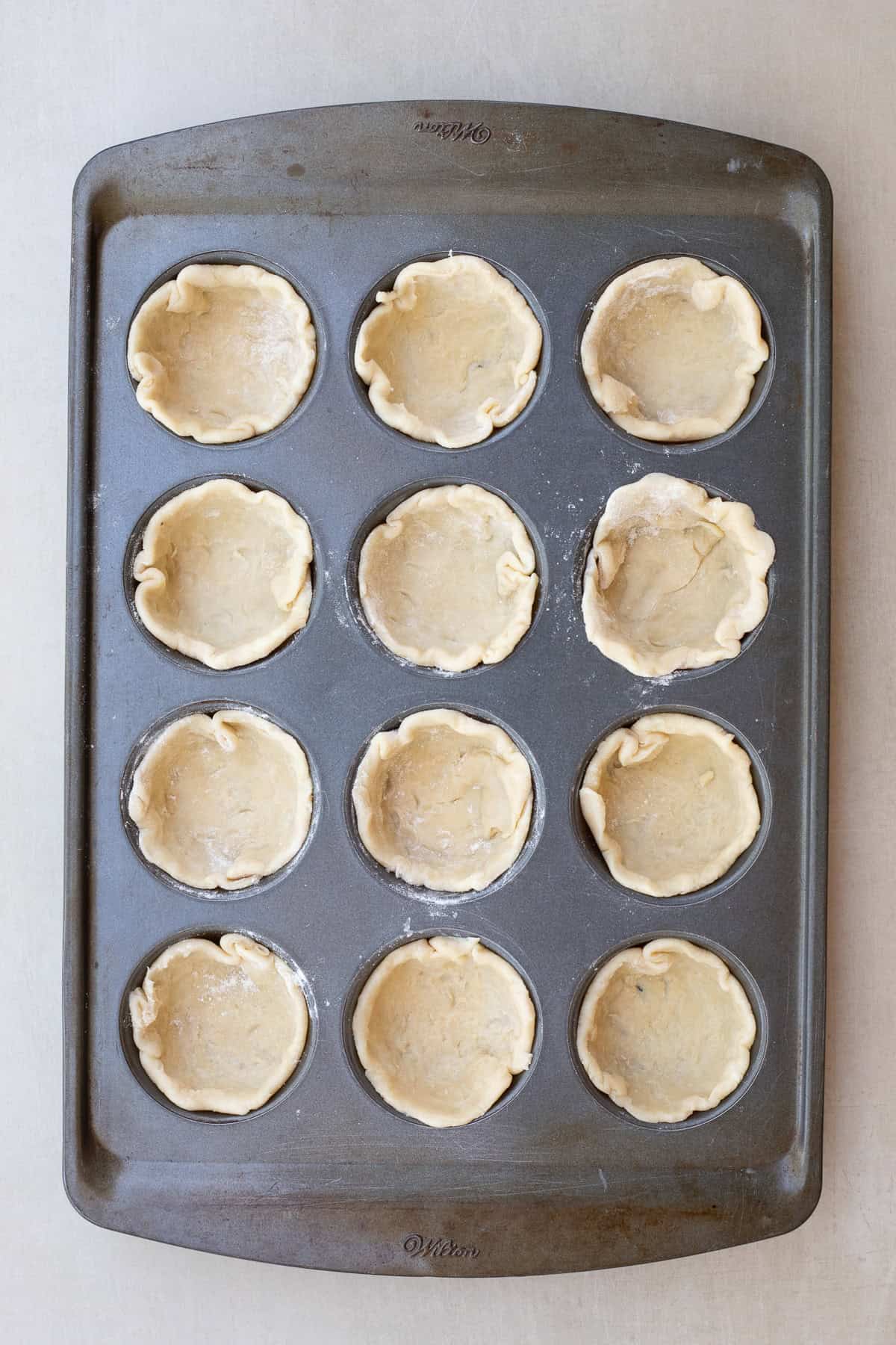 An easy mini quiche recipe with a buttery pie crust, and an egg filling. A muffin pan with all of the pie crust shaped into it.