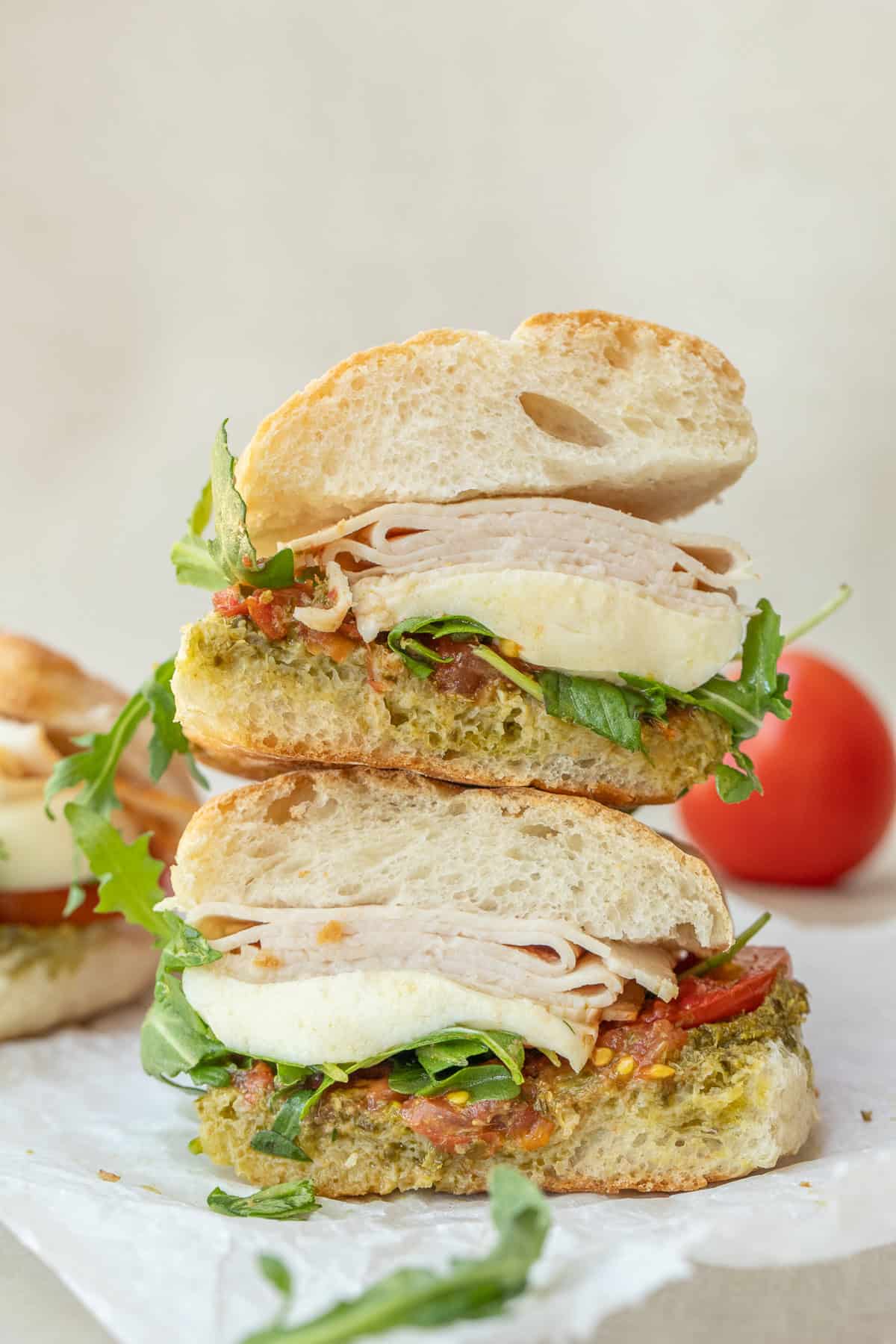 A chicken pesto sandwich is the perfect flavorful and delicious lunch. Soft and airy ciabatta bread, topped with homemade pesto, tomatoes, melty cheese, and deli chicken. Two pesto chicken sandwiches stacked on top of each other.