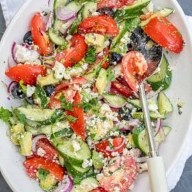 An avocado greek salad served in a salad bowl with a serving spoon.