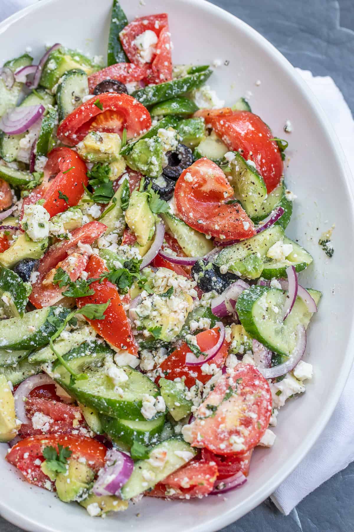 Fresh and flavorful avocado Greek salad with tomatoes, cucumbers, avocado, olives, red onion, and crumbled feta. A salad bowl with the avocado greek salad.