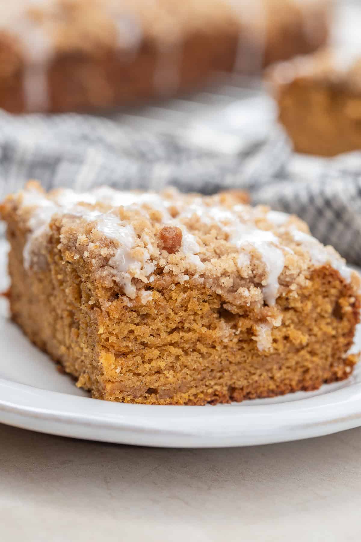 A slice of homemade pumpkin coffee cake with a moist pumpkin crumb, spiced streusel, and vanilla glaze. A slice of moist pumpkin coffee cake on a plate.