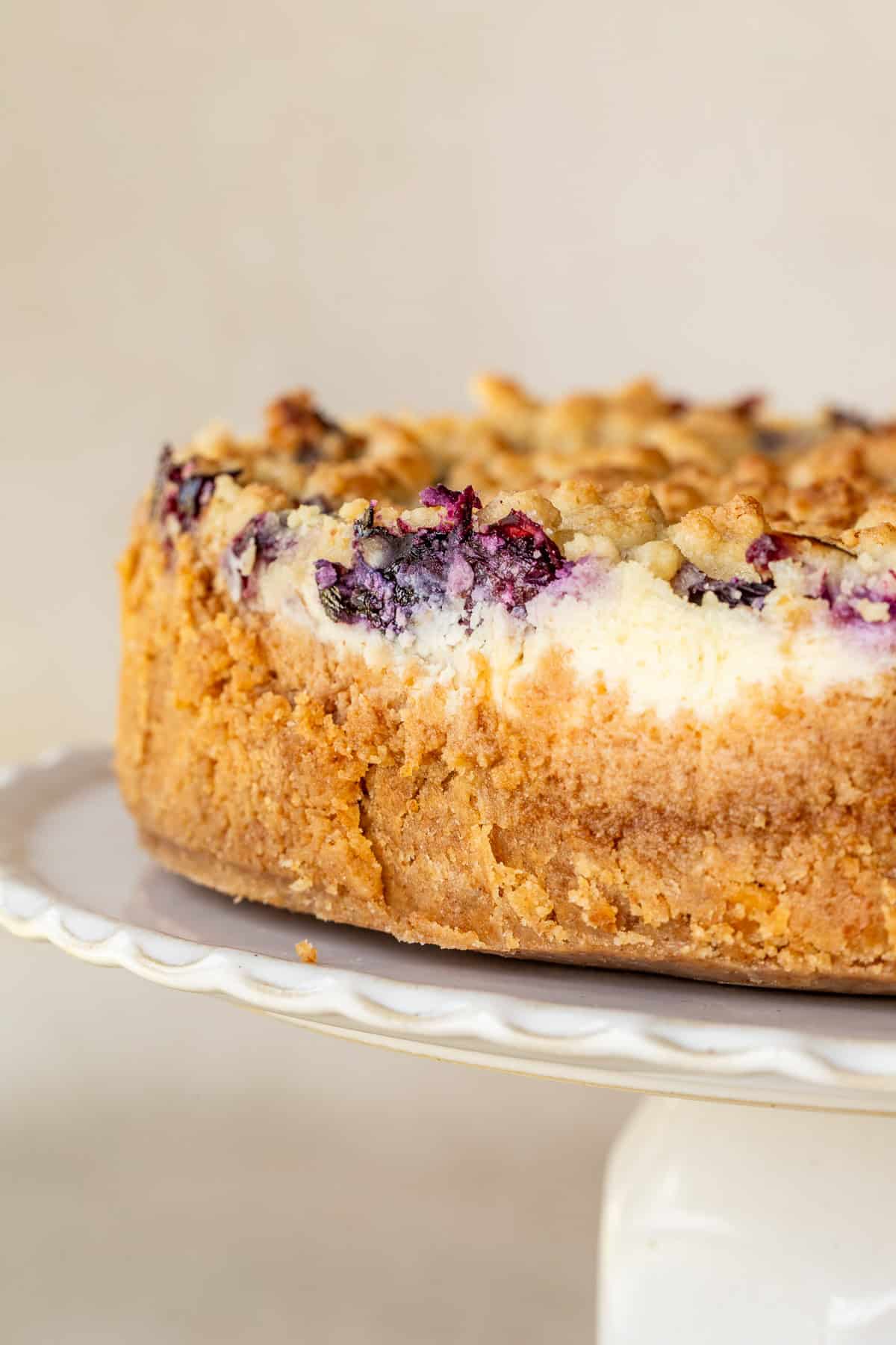 Homemade blueberry crumble cheesecake topped with sweet blueberries and golden buttery crumbs. A cake stand with the blueberry cheesecake.