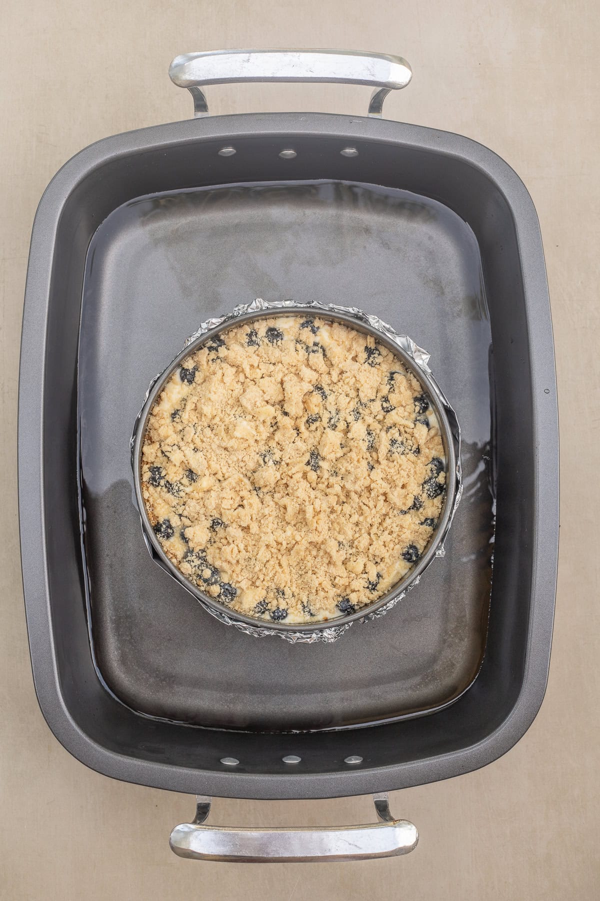Blueberry crumble cheesecake made with a creamy filling, sweet blueberry sauce, and crumb topping. A water bath made for the cheesecake using a large baking dish. The cheesecake is wrapped in foil to prevent any leaking.